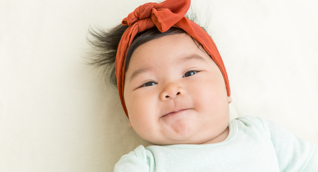 Smiling baby lying on a soft cream blanket, wearing a rust red bow headband and pale mint onesie, for a baby names article.