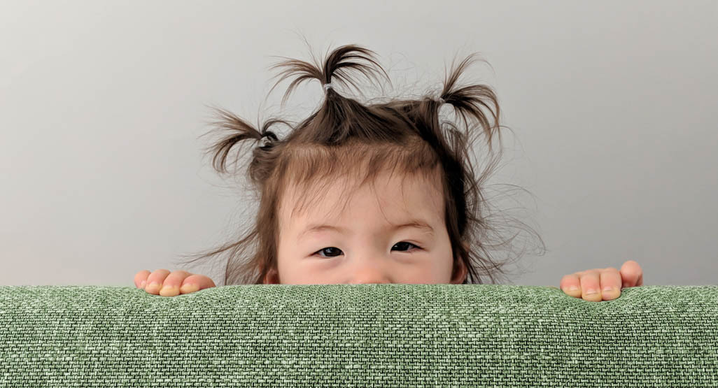 Young child with playful pigtails peeking over a green fabric couch, showing curious eyes and tiny hands against a soft gray background.
