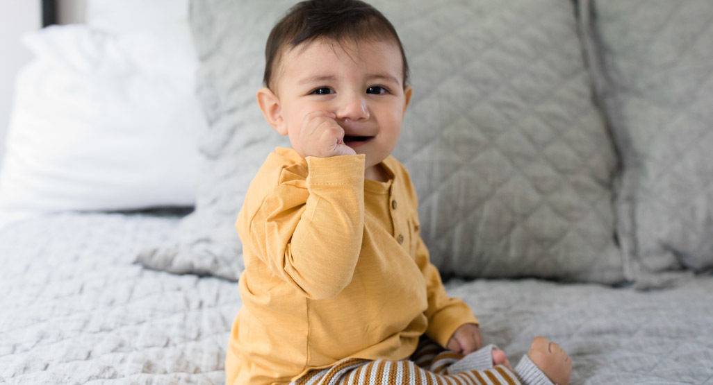 Smiling baby in a mustard yellow shirt sitting on a bed with gray pillows, featured for a Hebrew and Jewish baby names article.