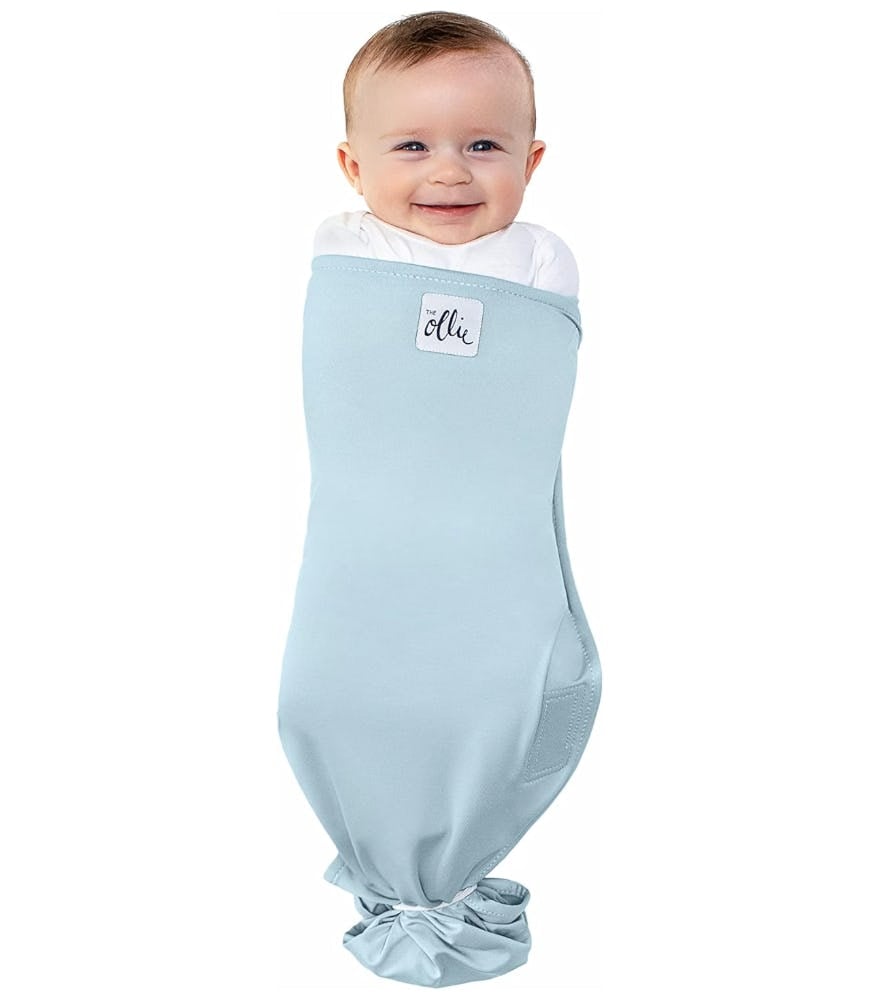 Smiling baby wrapped in a light blue Ollie swaddle on a white background, showcasing a soft newborn sleep sack for a baby registry.