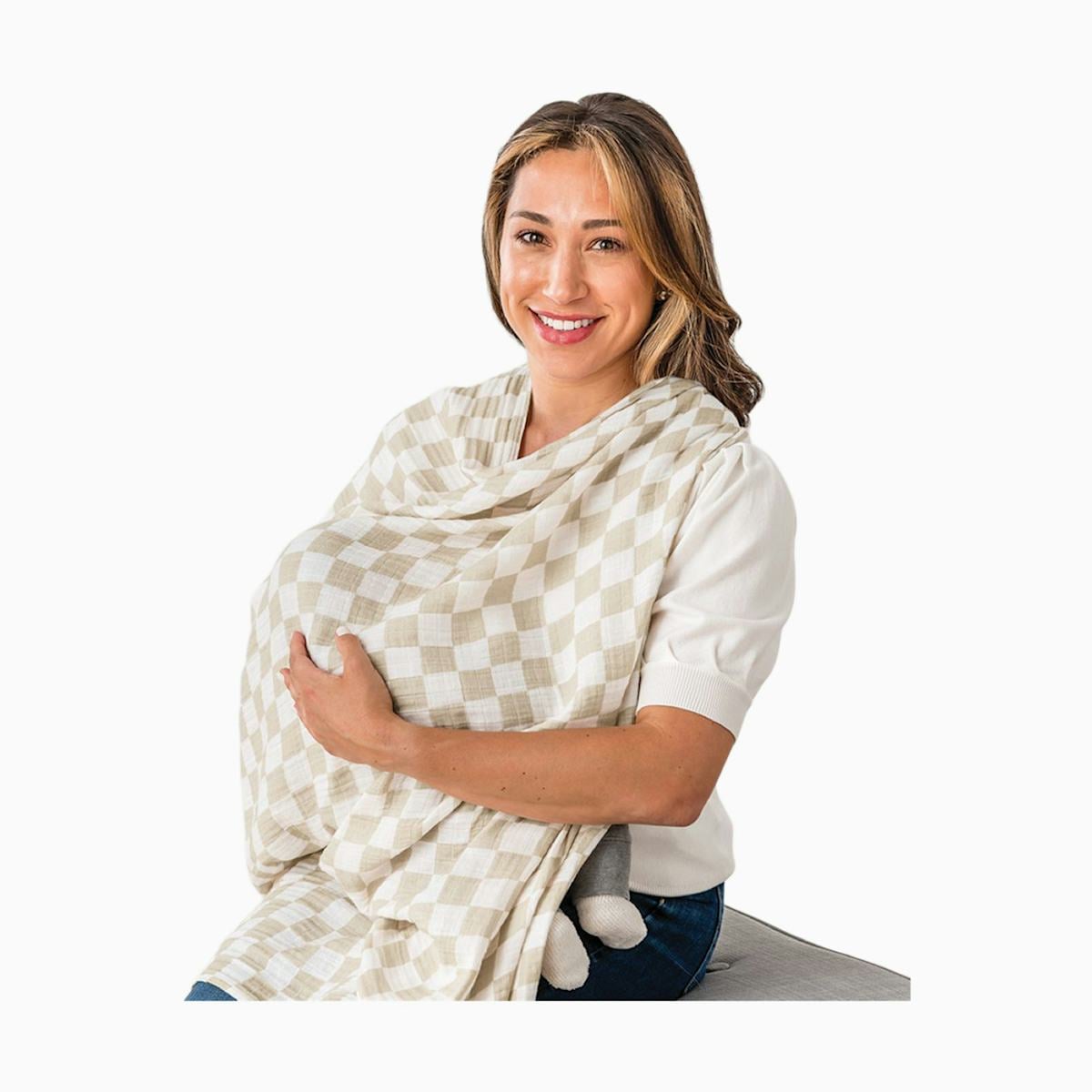 Smiling mother breastfeeding a baby under a beige checkered nursing cover on a white studio background