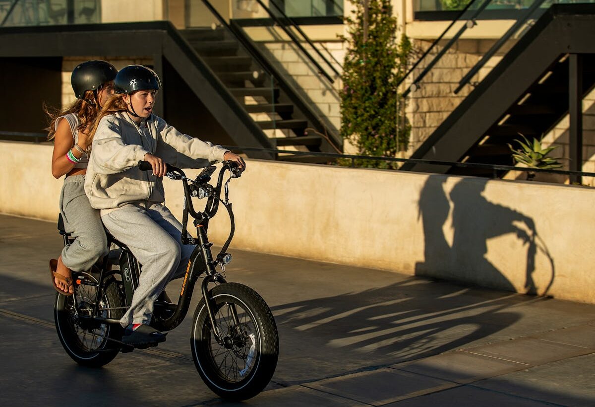 Two helmeted young riders share a black electric bike on a sunlit urban street, casting a long shadow beside modern apartment buildings.