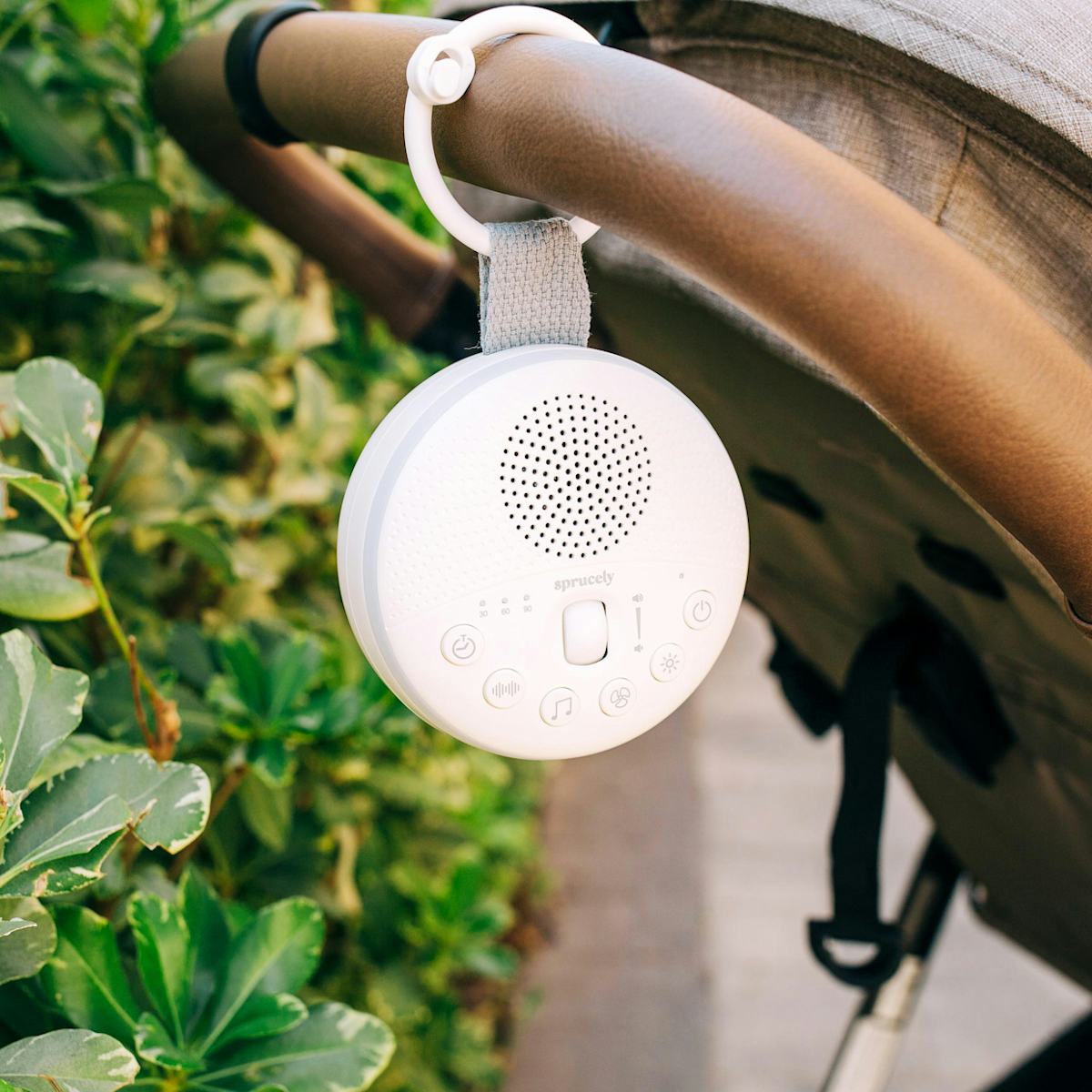 White Sprucely travel sound machine clipped to a stroller handle outdoors, surrounded by greenery for on-the-go baby naps.