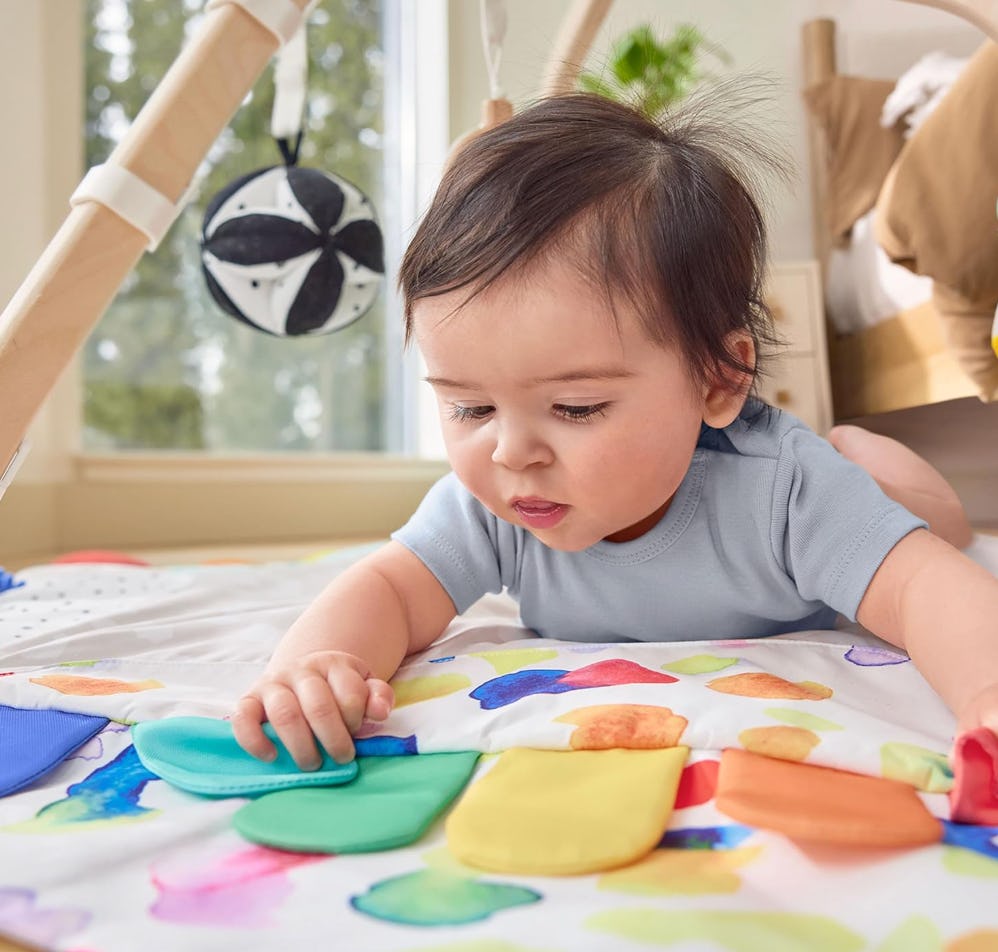 Baby doing tummy time on a colorful play mat, reaching for soft sensory toys beneath a hanging black-and-white play gym mobile.