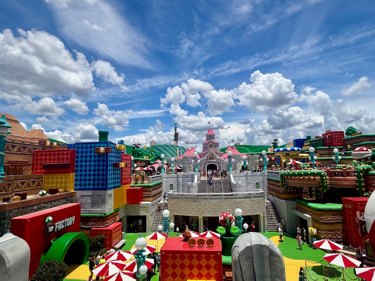 Colorful Super Nintendo World at Universal Epic Universe with Peach’s Castle, Mario-themed buildings, crowds, umbrellas, and a bright blue sky.
