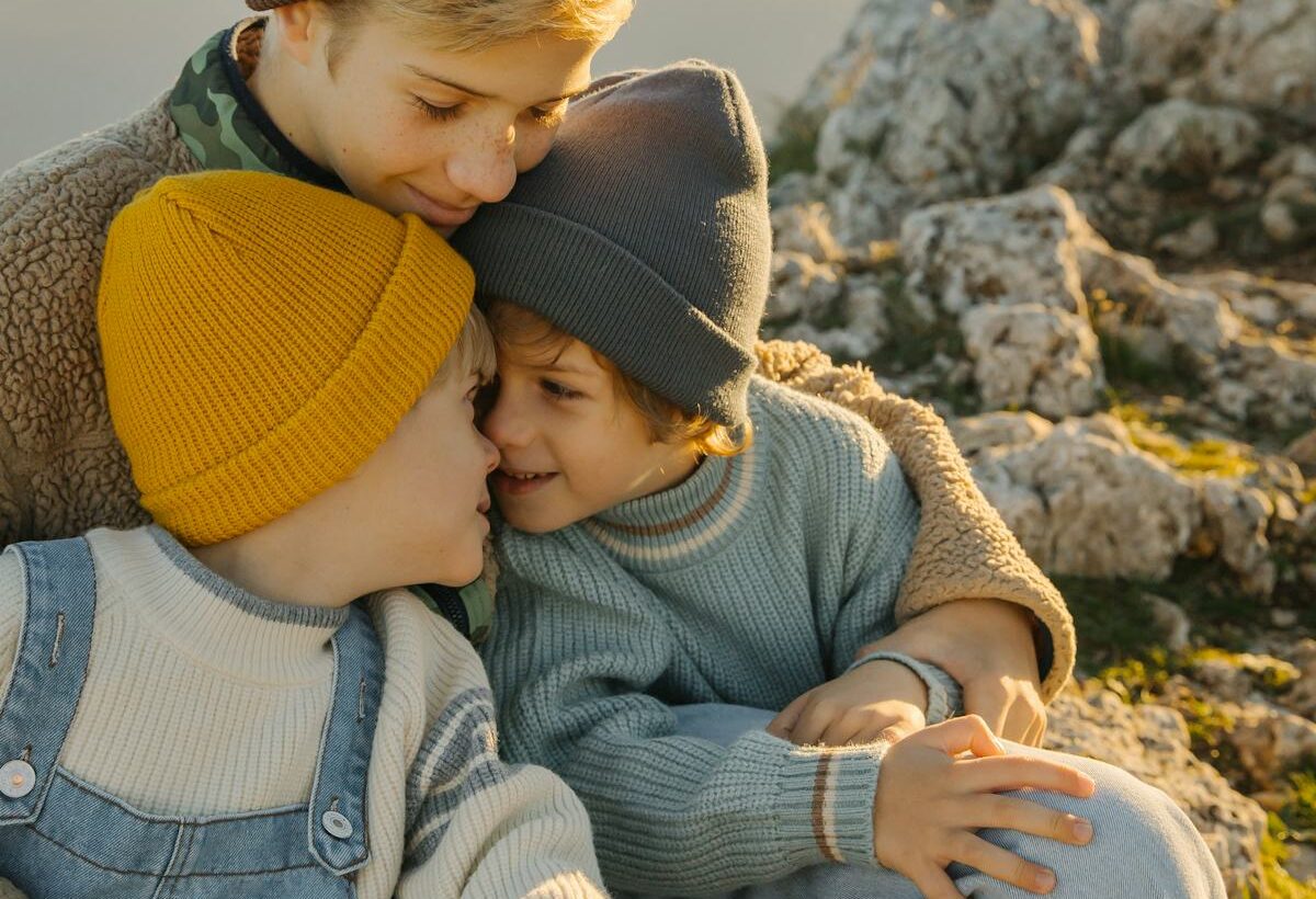 Three children in knit hats cuddle together on a rocky mountain at sunset, showing sibling closeness and family bonding outdoors.
