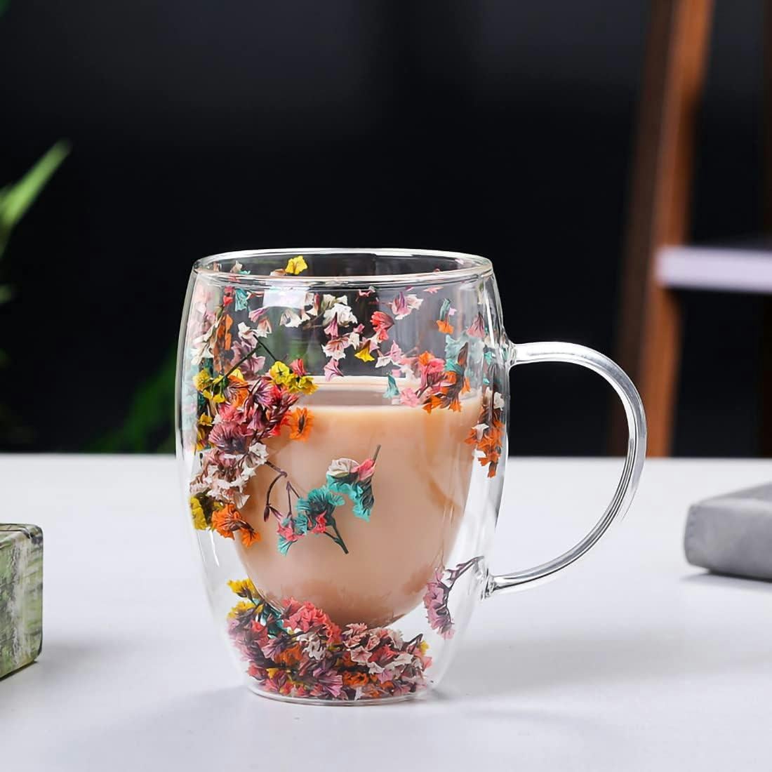 Double-walled glass coffee mug with colorful dried flowers between the glass layers, filled with a creamy latte on a white table.