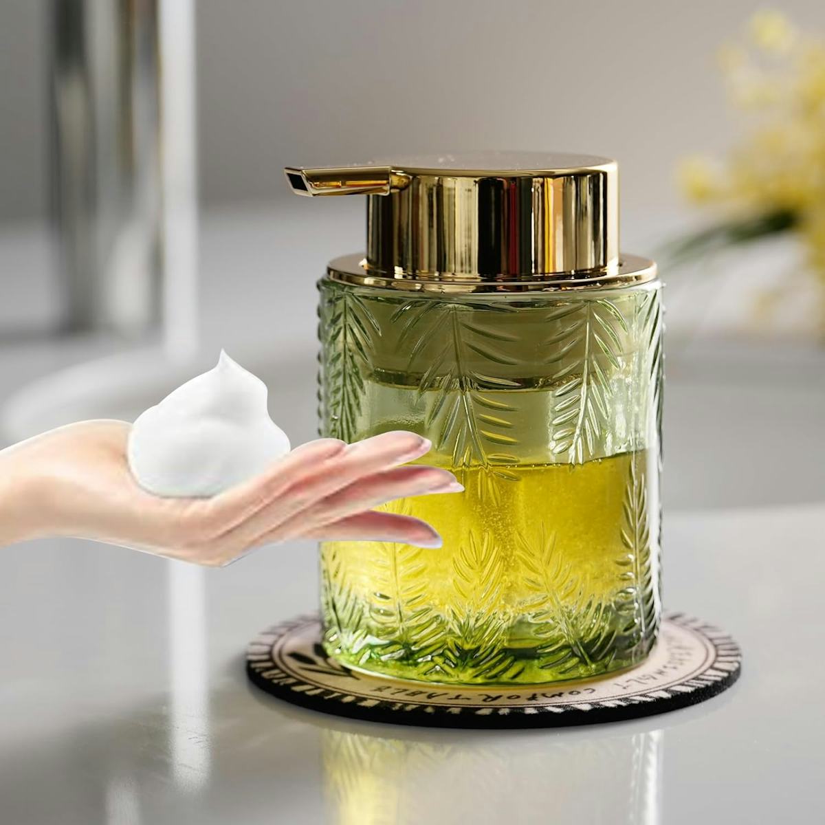 Green ribbed glass foaming soap dispenser with gold pump beside a hand holding white foam on a bathroom counter