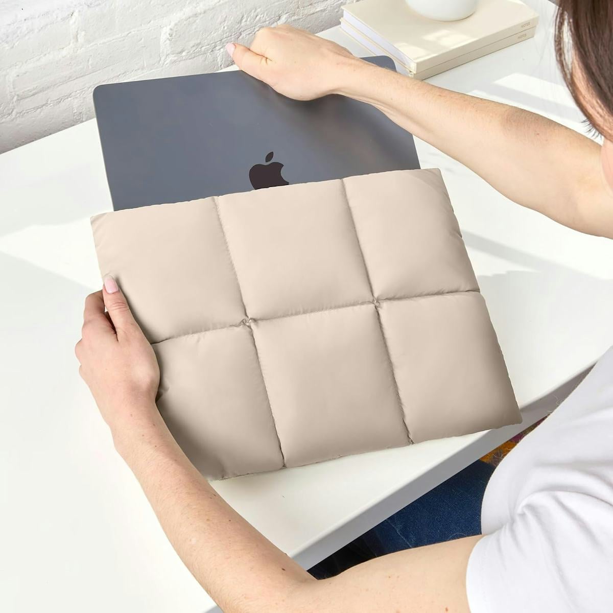 Beige quilted puffy laptop sleeve protecting a gray MacBook on a white desk as a person slides the computer inside.