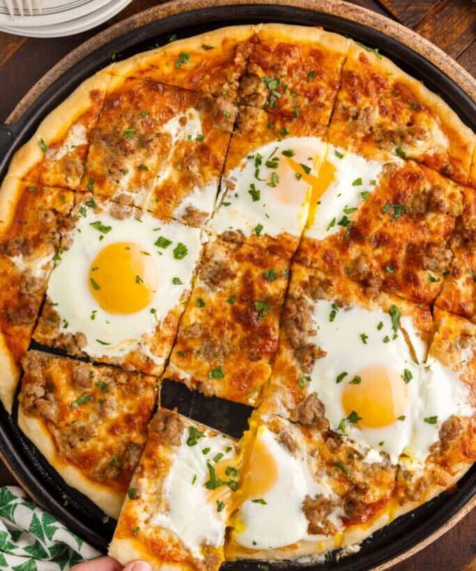 Breakfast pizza topped with eggs, sausage, melted cheese, tomato sauce, and parsley, sliced on a black pan over a wooden table.