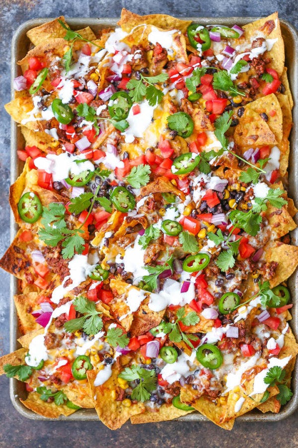 Sheet pan nachos topped with melted cheese, ground beef, black beans, corn, diced tomatoes, red onion, jalapenos, cilantro, and crema.