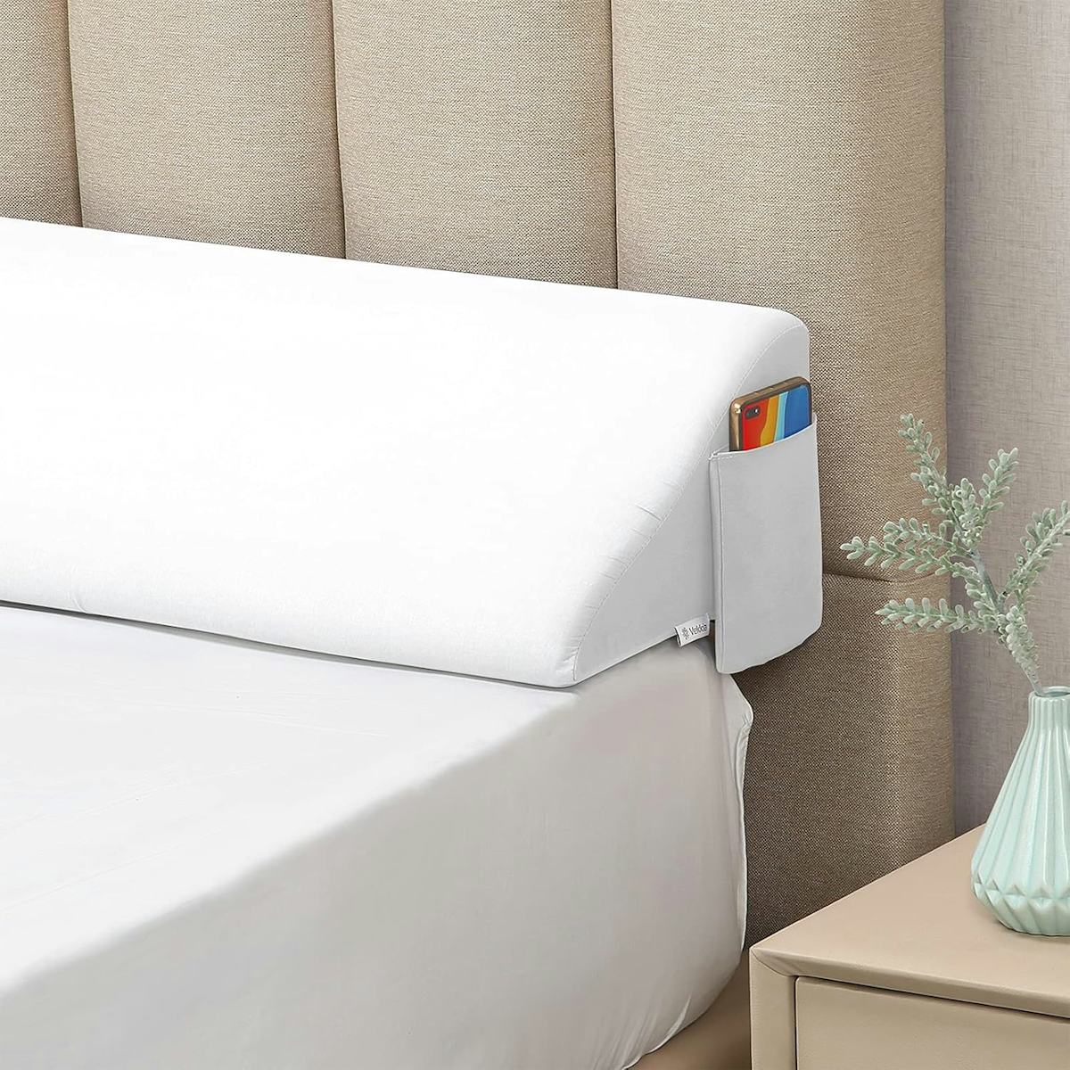 White wedge bed gap pillow with side pocket holding a phone between mattress and beige upholstered headboard