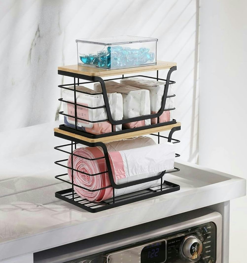 Two-tier black wire and bamboo countertop organizer holding trash bags, wipes, and a clear box on a laundry room counter