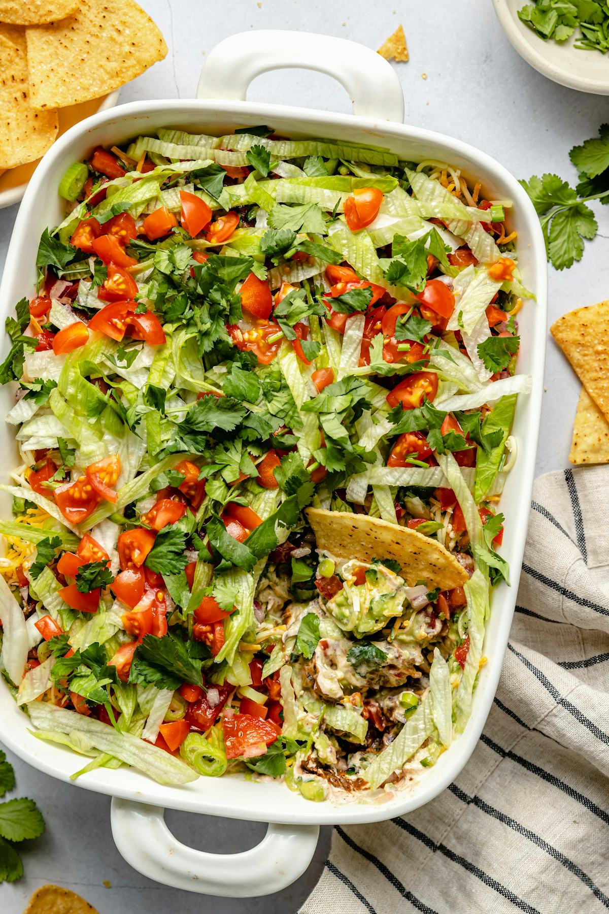 Seven-layer dip topped with shredded lettuce, diced tomatoes, cilantro, and tortilla chips in a white baking dish.