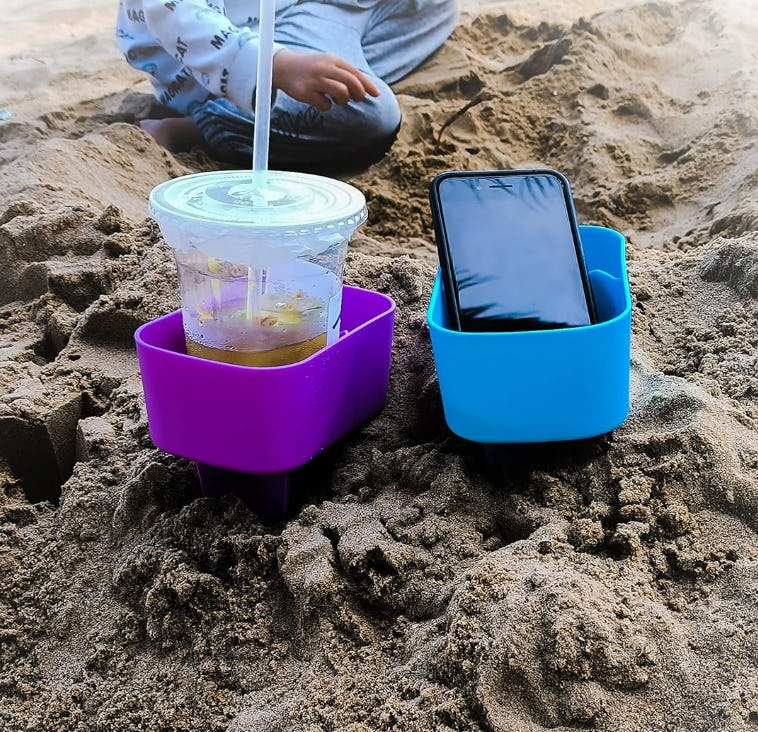 Purple and blue beach cup holders stuck in sand, holding an iced drink with straw and a smartphone during a beach outing.