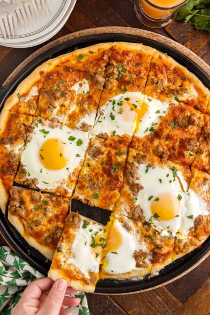 Breakfast pizza with baked eggs, sausage, melted cheese, tomato sauce, and fresh herbs sliced on a black pan over a wooden table.
