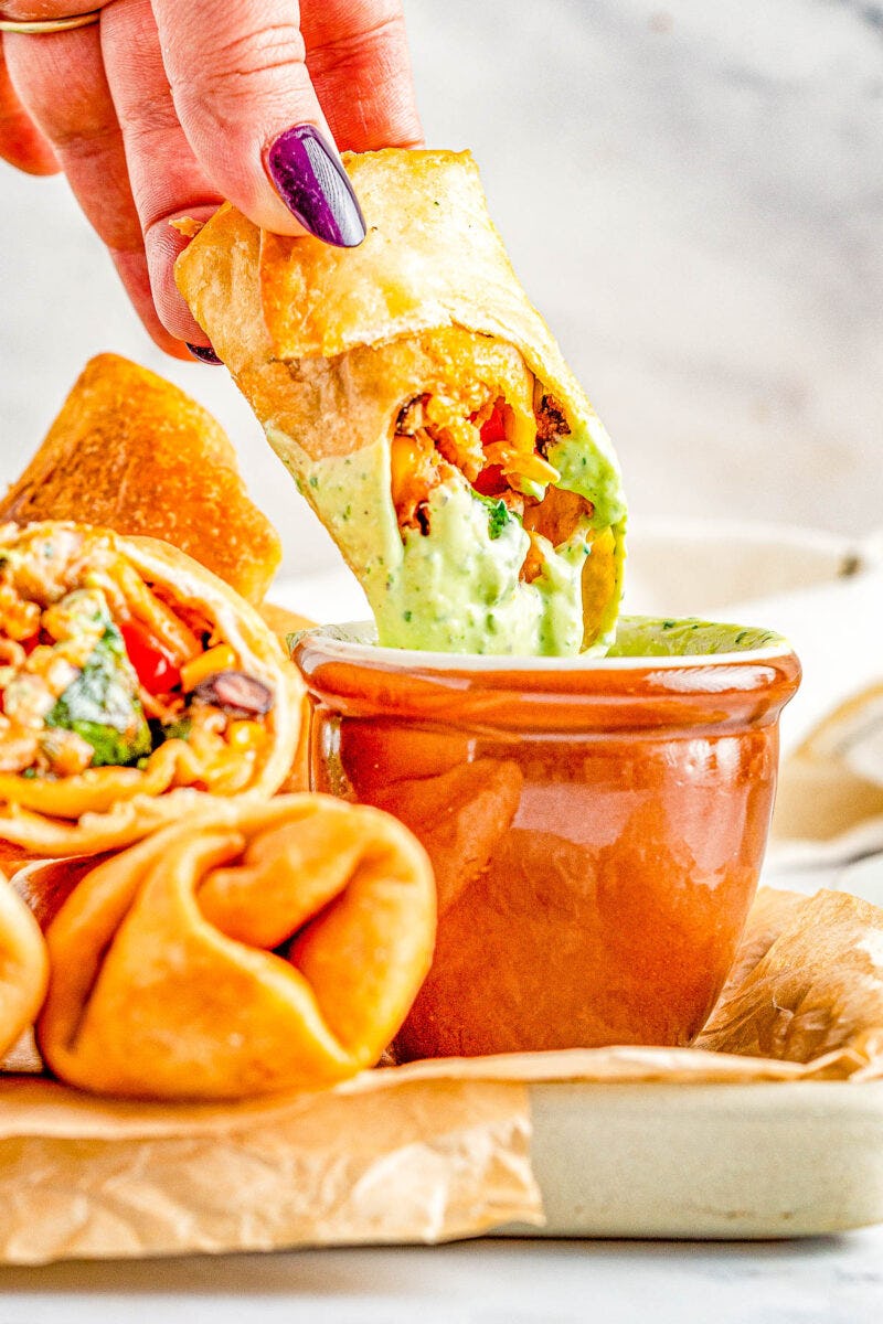 Hand dipping a crispy Southwestern egg roll into creamy green avocado sauce beside more fried rolls on a tray.