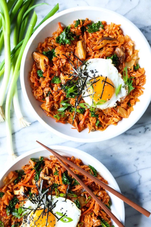 Two white bowls of kimchi fried rice topped with sunny-side-up eggs, seaweed, sesame seeds, scallions, and chopsticks on marble.