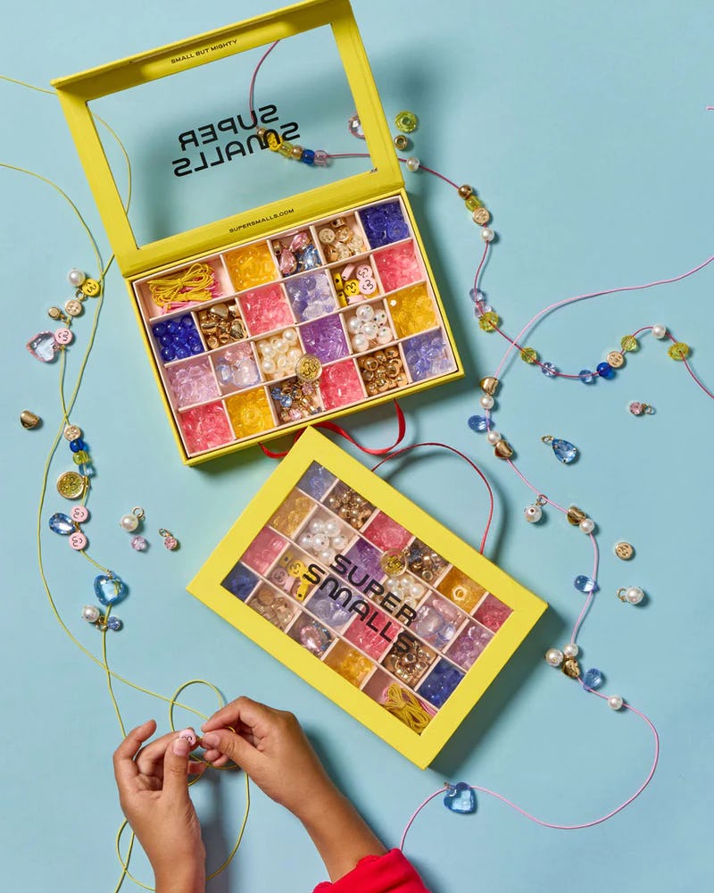 Super Smalls kids bead kit with colorful charms, pearls, strings, and yellow organizer boxes on a blue craft table.