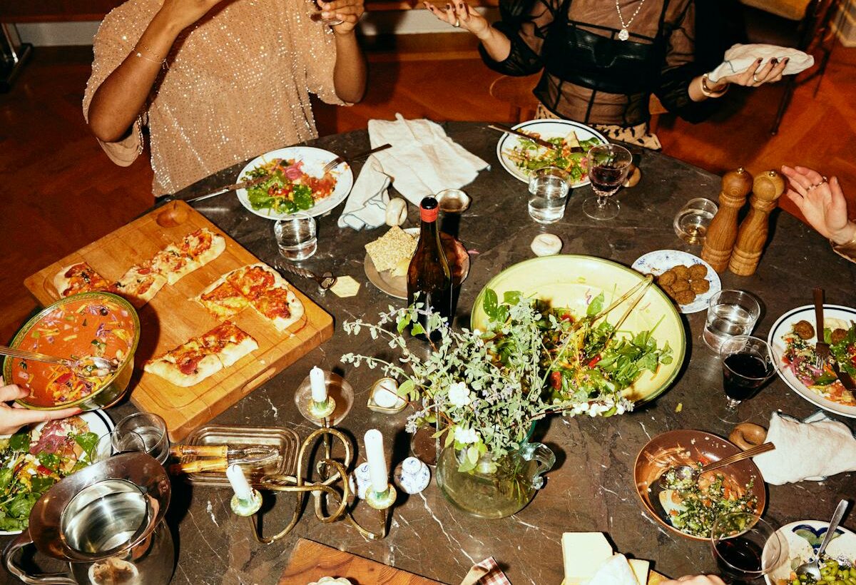 Friends gather around a dinner table filled with salads, bread, cheese, wine, and shared dishes for a lively potluck meal.