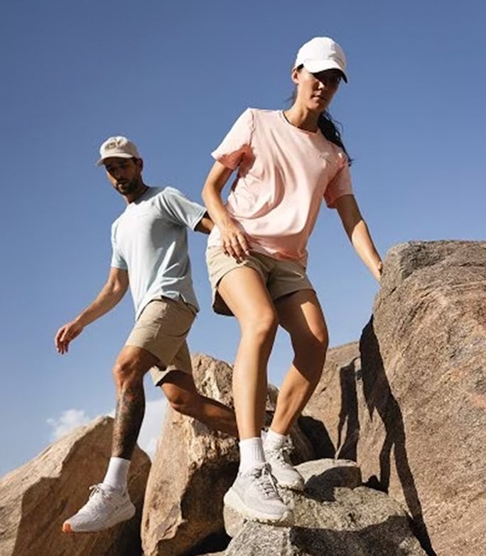 Two hikers in caps and light athletic clothing climb down sunlit desert rocks under a clear blue sky during a warm outdoor adventure.