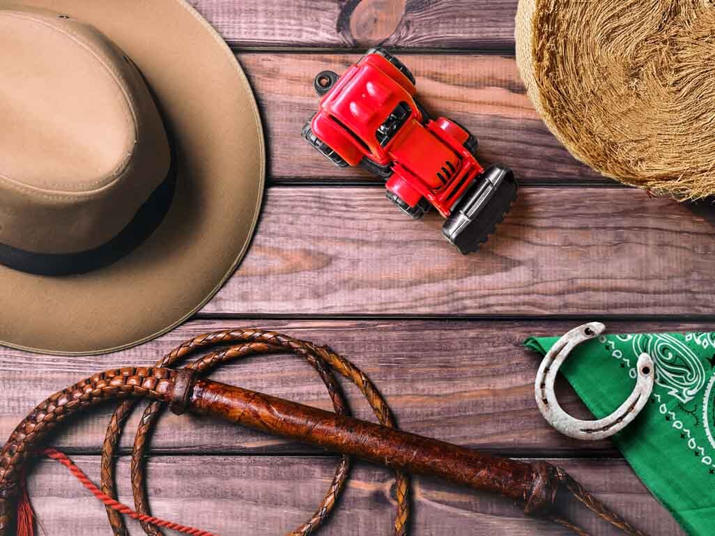 Western baby name themed flat lay with cowboy hat, toy red tractor, rope, hay bale, horseshoe, and green bandana on rustic wood.