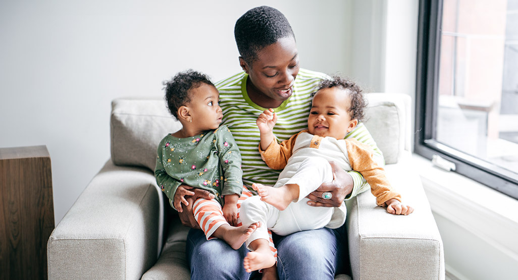 Parent sitting in an armchair by a bright window, holding two young children and smiling during a warm family moment.