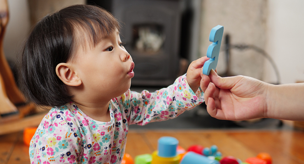 A curious toddler reaches toward a blue question mark toy held by an adult, suggesting baby name choices and spelling questions.