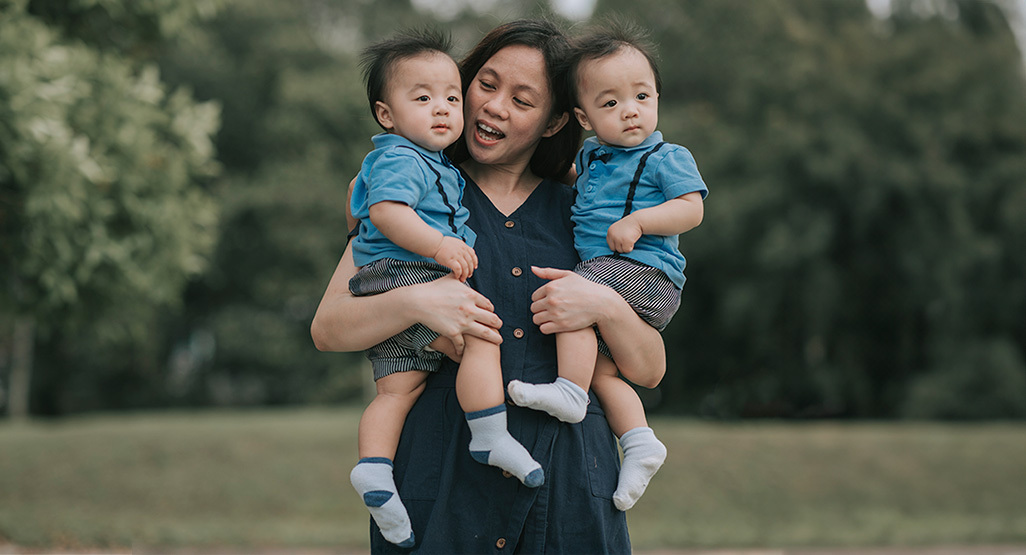 Mother holding twin babies in matching blue outfits outdoors, smiling together in a soft-focus park setting.