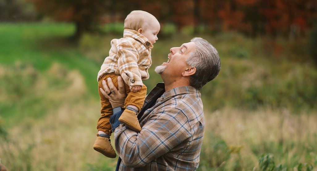 Smiling grandfather lifting a baby in a plaid jacket outdoors in a grassy autumn field, sharing a warm family moment.