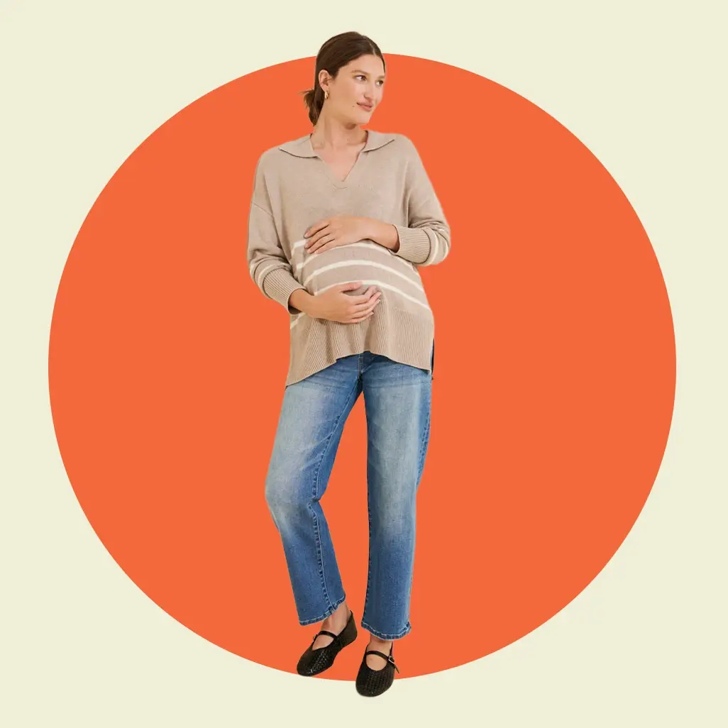 Pregnant woman wearing a beige sweater, blue maternity jeans, and black flats posing in front of an orange circle.