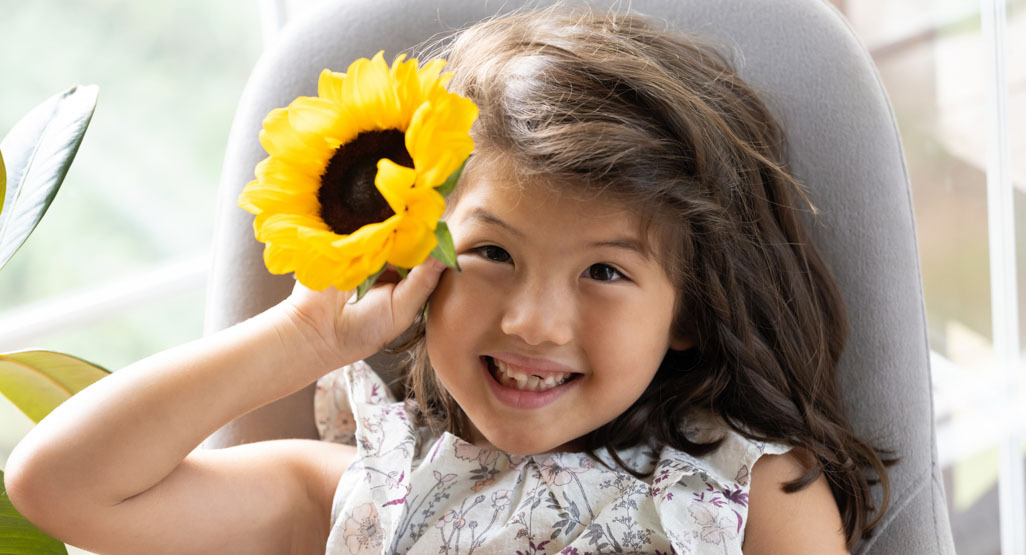 Smiling young girl holding a bright yellow sunflower over one eye while sitting indoors in a soft gray chair near a sunny window.