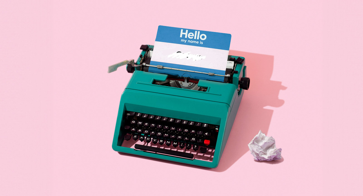 Turquoise typewriter on a pink background holding a Hello my name is tag, with a crumpled paper ball nearby.
