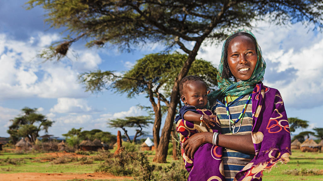 African mother holding her baby in a colorful wrap beneath acacia trees in a rural village landscape with huts and blue sky.