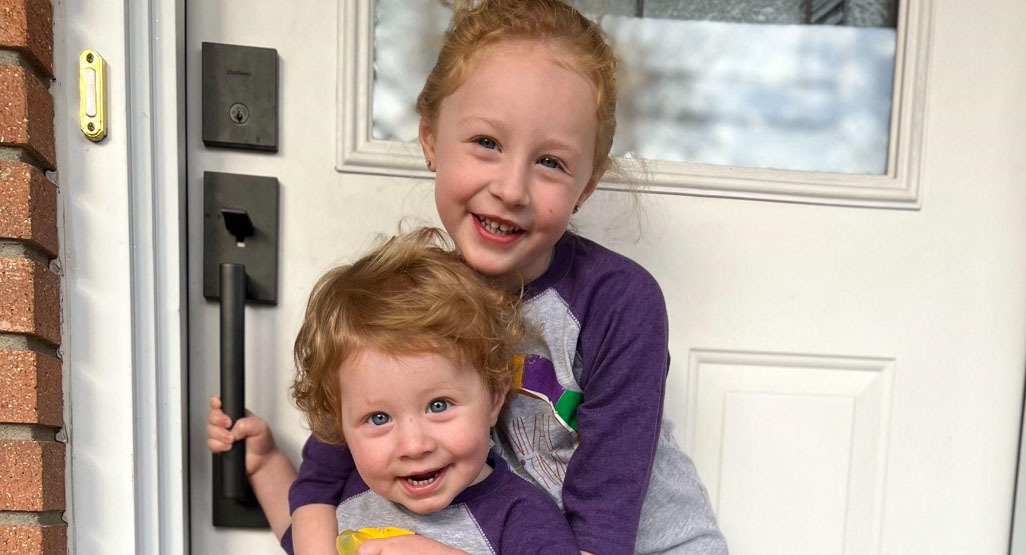 Smiling young siblings in matching purple shirts hug at a front door, with the toddler boy holding the handle and his older sister behind him.