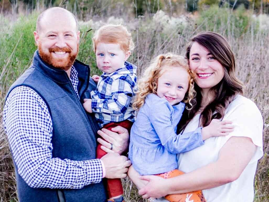 Smiling family of four posing outdoors, with parents holding a young girl and little boy in a grassy field.