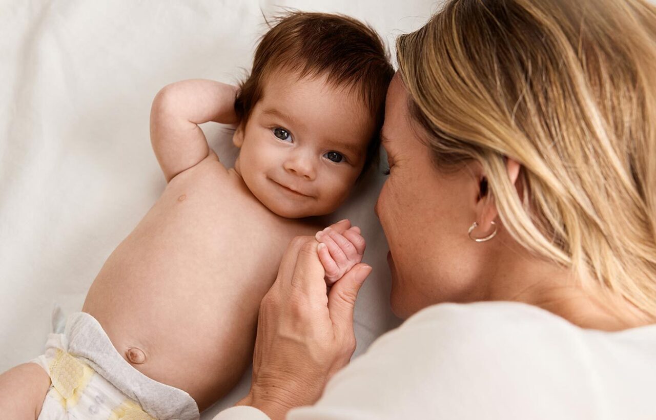 Mother lying beside smiling baby in a diaper on white bedding, capturing a tender nursery moment for baby registry essentials.