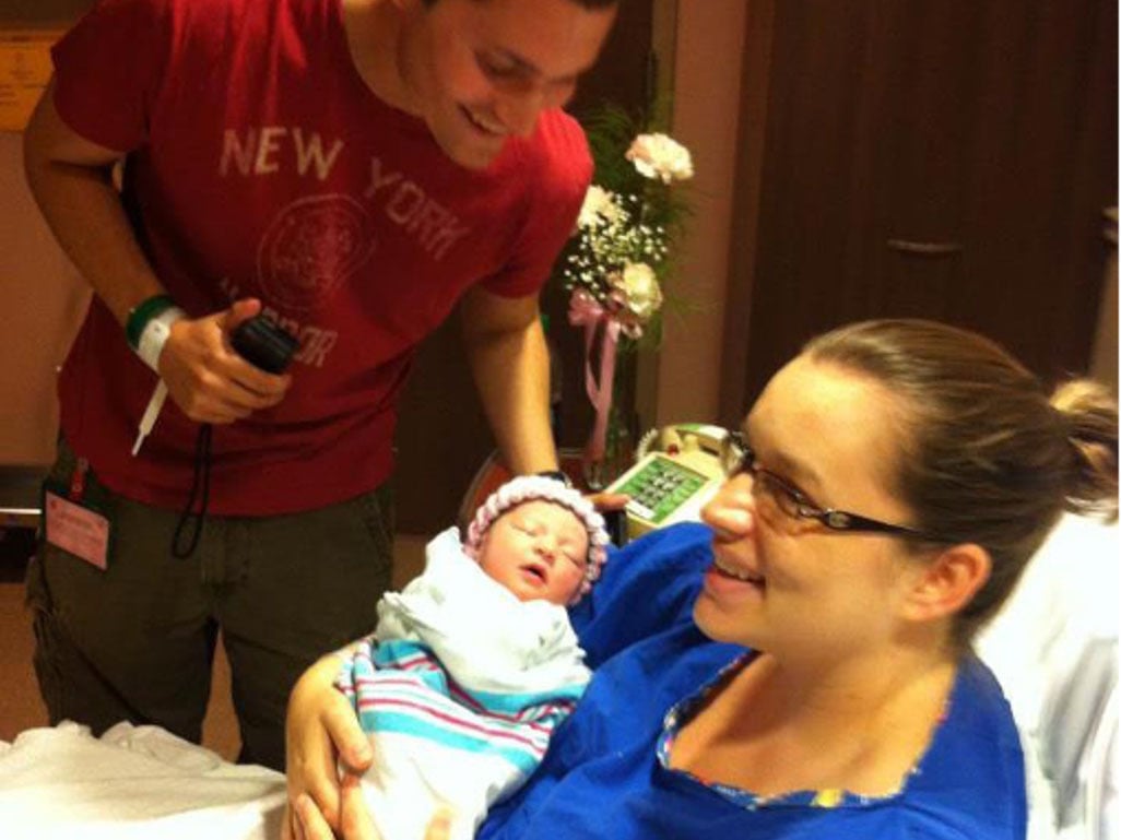 New parents smile over a swaddled newborn baby in a hospital room shortly after birth, with medical equipment and flowers nearby.
