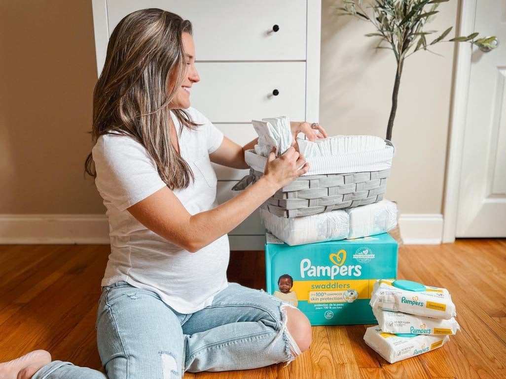 Pregnant woman organizing Pampers diapers and wipes in a nursery while preparing baby registry essentials on a hardwood floor.
