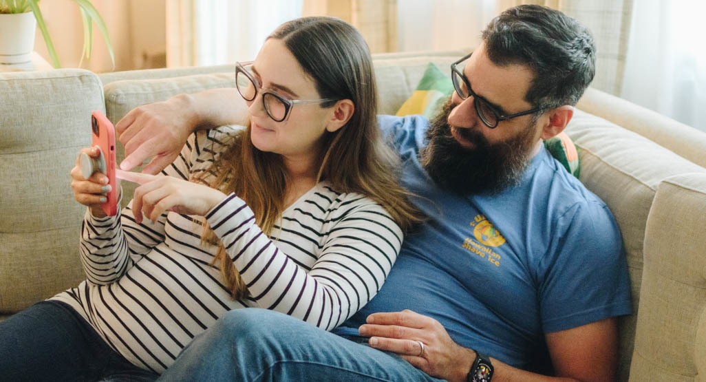 Expectant couple relaxing on a couch and looking at a smartphone while planning a baby registry at home.