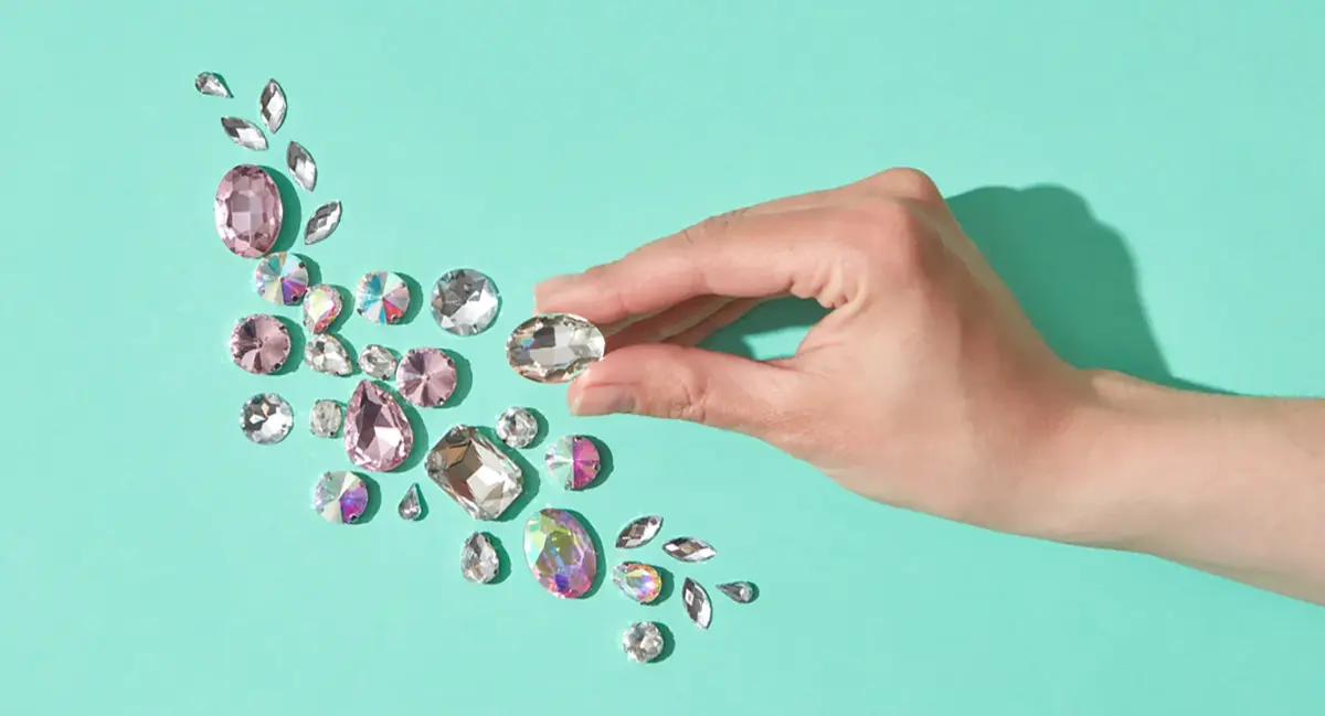 A hand arranges sparkling pink and clear rhinestones on a turquoise background, suggesting rare gems and uncommon baby girl names.