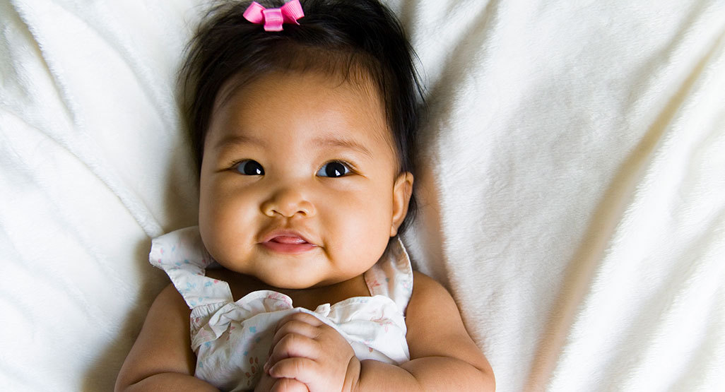 Smiling baby girl lying on a soft white blanket, wearing a light ruffled outfit and a small pink bow in her dark hair.