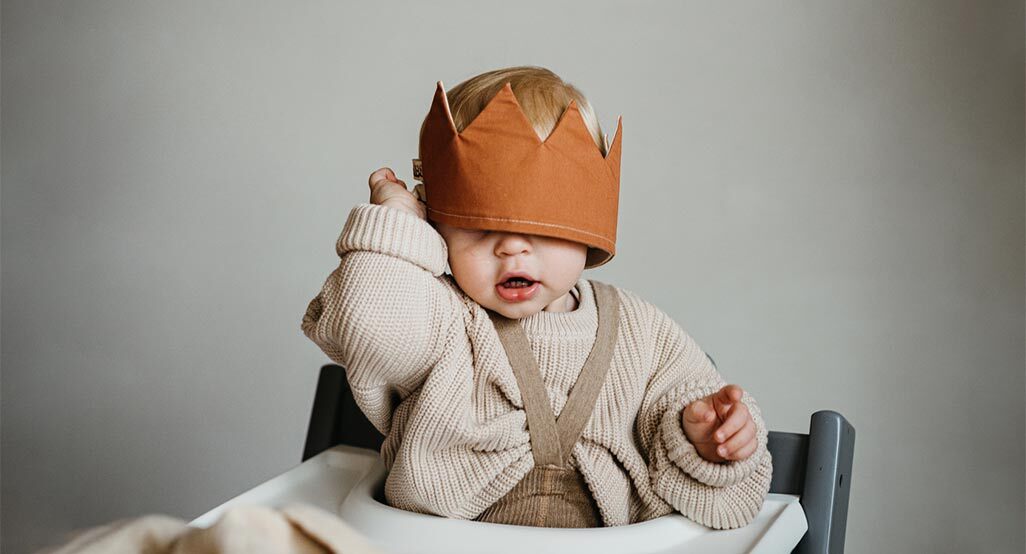 Baby wearing a brown paper crown sits in a high chair, dressed in a cream knit sweater, evoking a playful royal baby names theme.