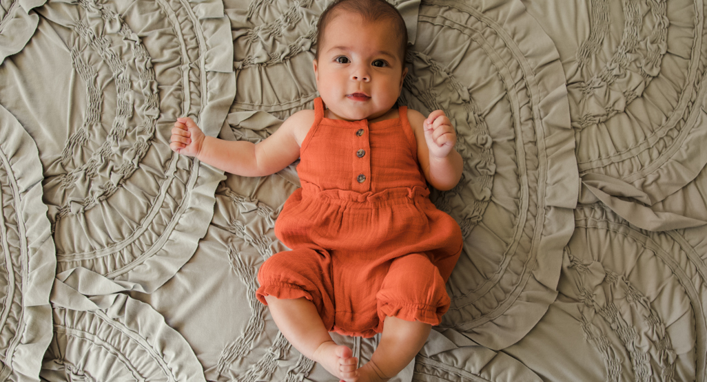 Smiling baby in a rust-orange romper lying on a textured gray blanket, ideal image for newborn care and baby name articles.