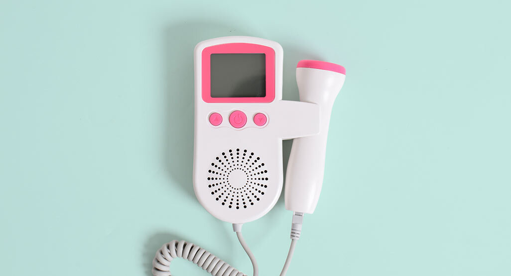 White and pink handheld fetal doppler monitor with probe on a mint green background for home pregnancy heartbeat checks.