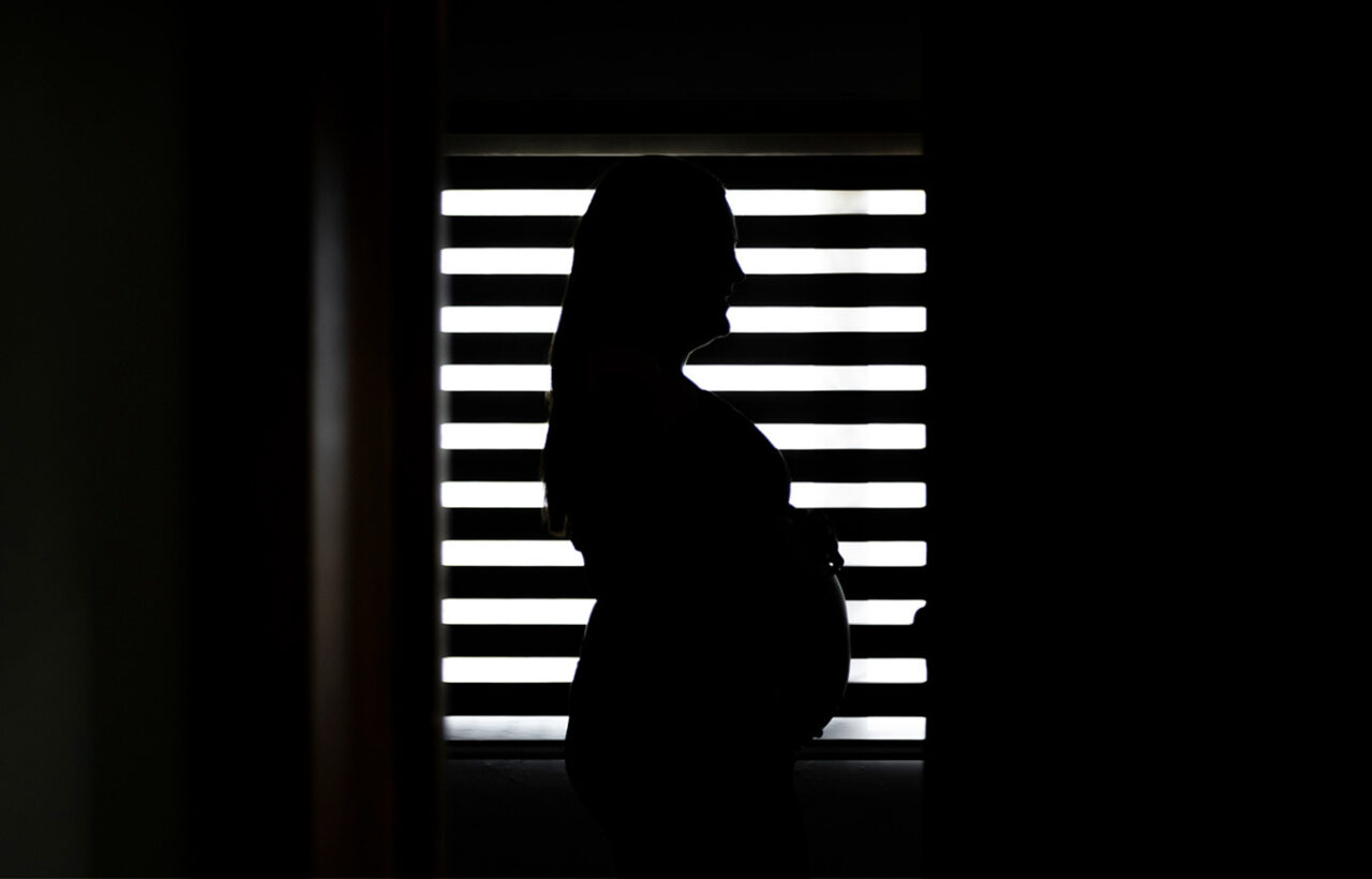 Silhouette of a pregnant woman standing in profile before bright window blinds in a dark room, suggesting prenatal care and pregnancy concerns.