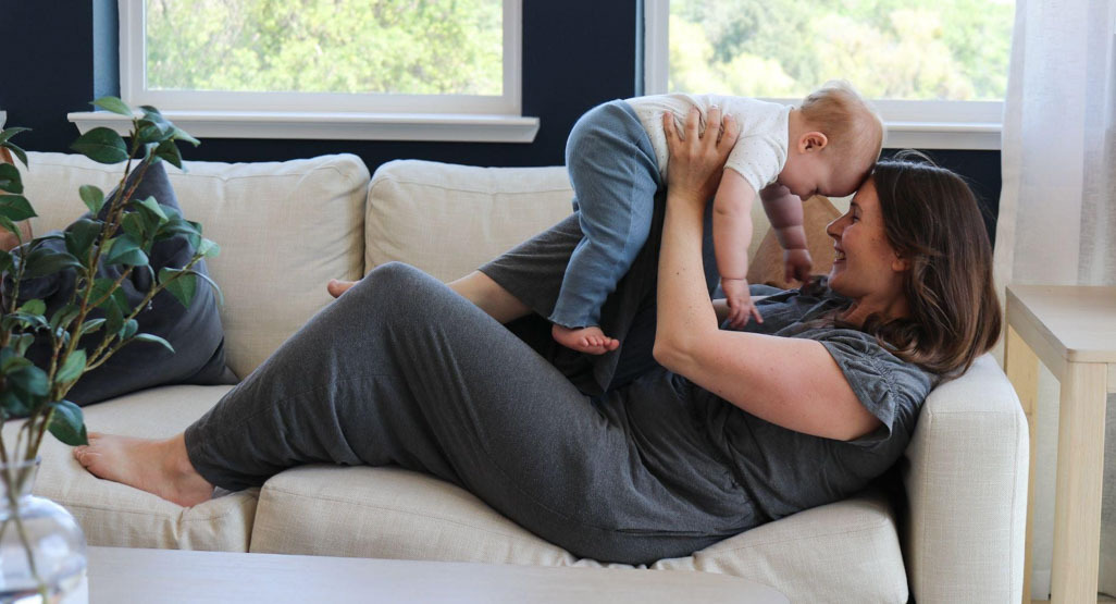 Mother lying on a couch lifts and smiles at her baby in a bright living room, suggesting nurturing care and infant wellness at home.