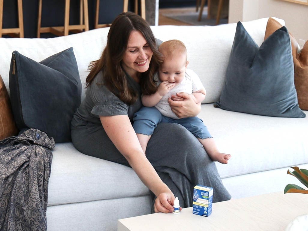 Mother sitting on a couch with her baby, reaching for vitamin D drops and packaging on a coffee table at home.