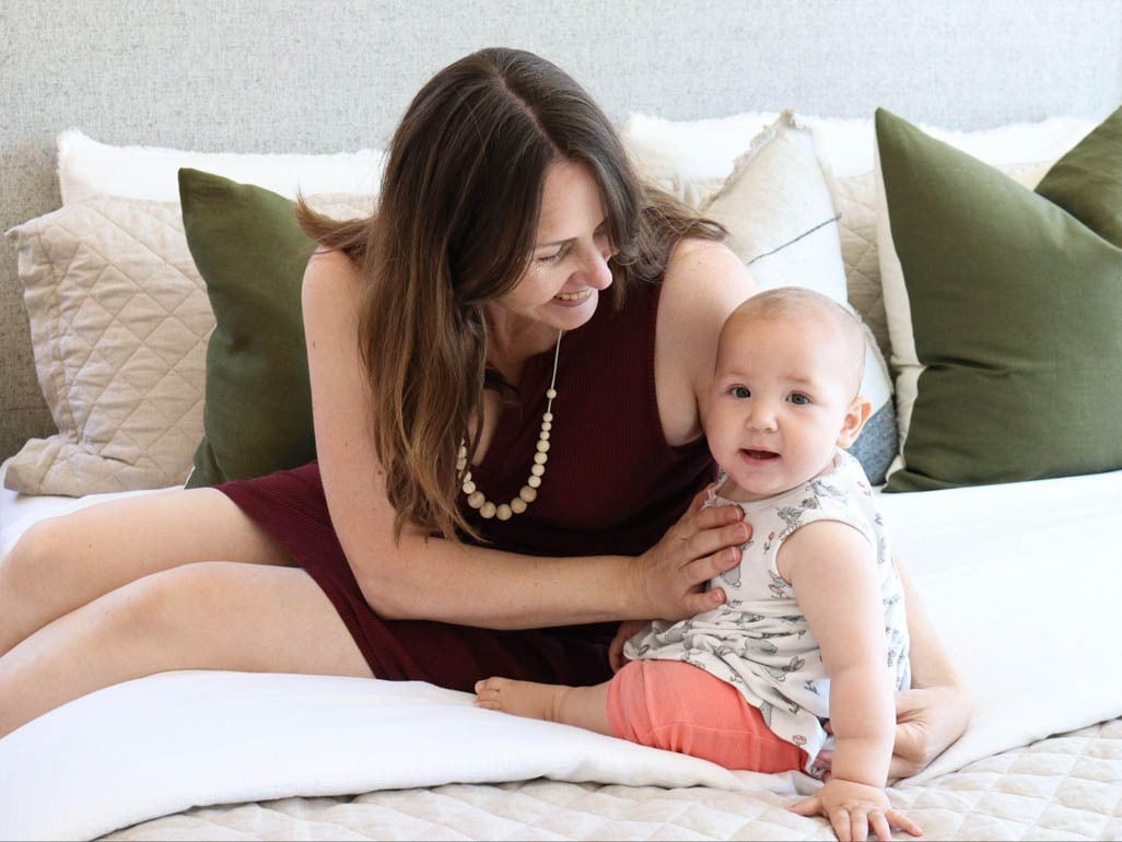 Mother smiling at her baby while they relax on a bed with pillows, a warm parenting image for infant health and vitamin D guidance.