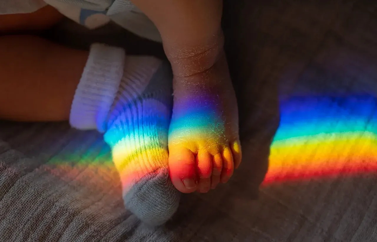 Baby feet resting on a soft blanket with a vivid rainbow of light across the toes and sock, symbolizing a rainbow baby.